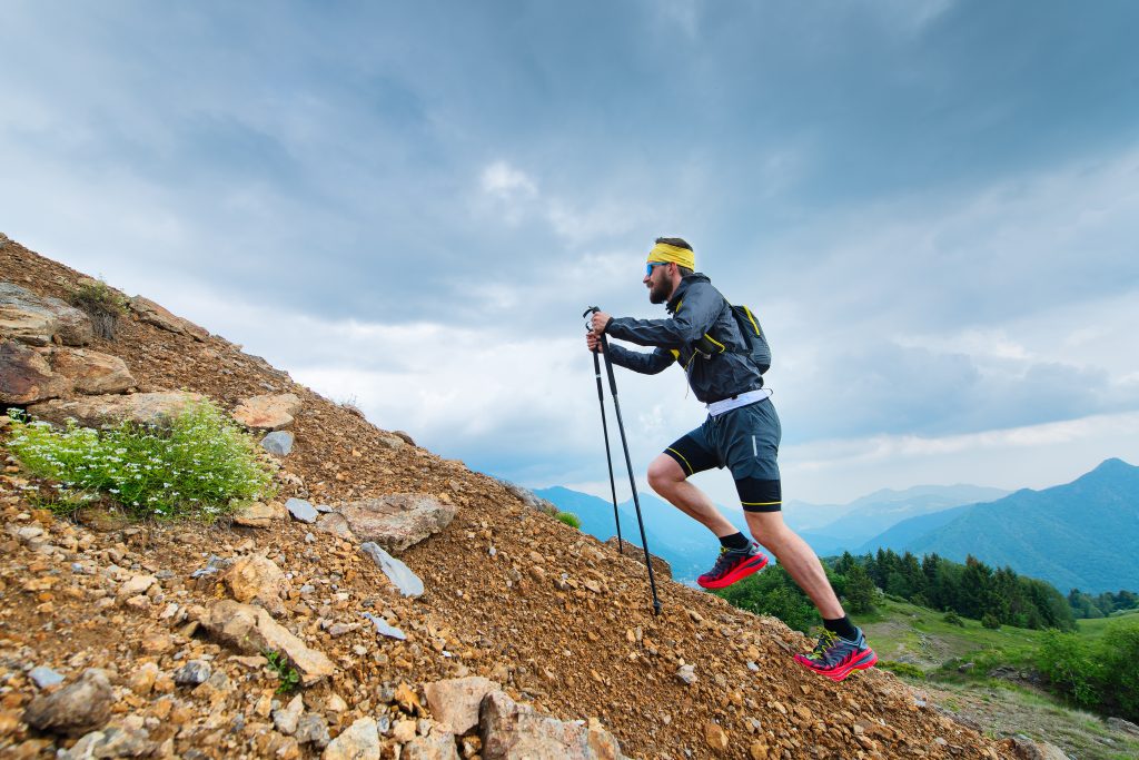 Un coureur de trail monte une pente raide en s'aidant de ses bâtons.