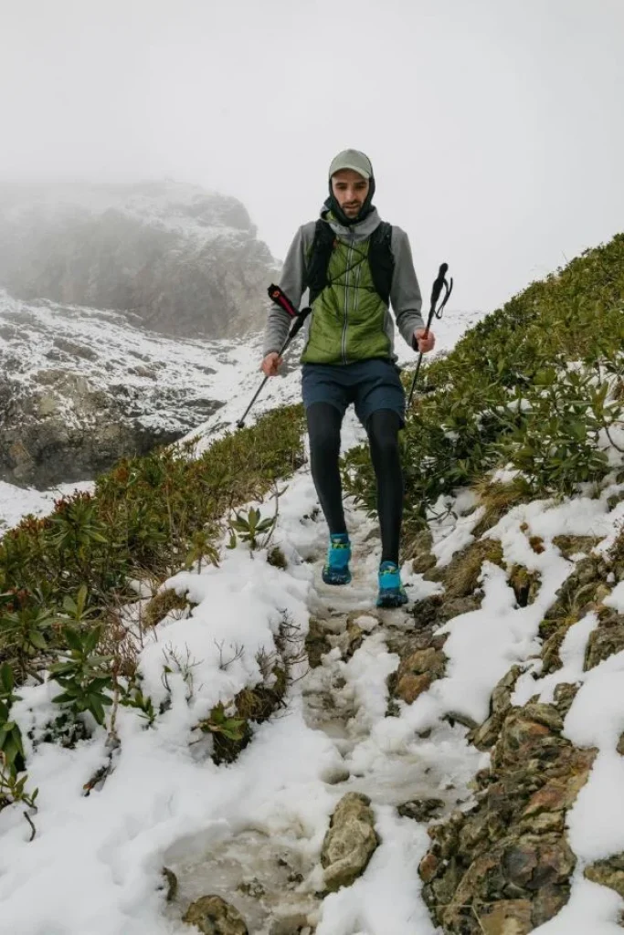 Un homme court sur un sentier de montagne enneigé avec un équipement de trail chaud et des bâtons.